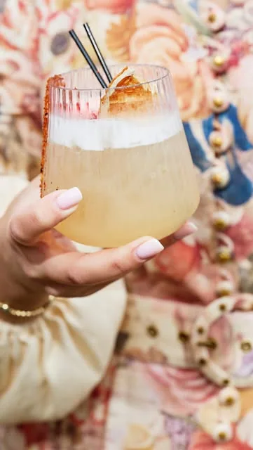 A guest holds a ribbed glass containing a pale cocktail topped with foam, dried citrus and fine chilli threads, finished with a spiced rim.
