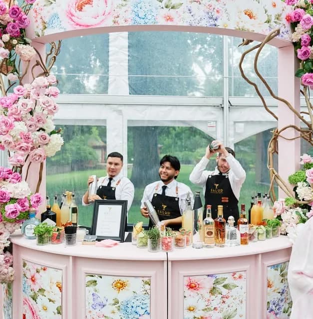 Three Salud bartenders in branded black aprons mixing drinks behind a curved bar clad in floral print, framed by pink and white roses at a garden marquee.