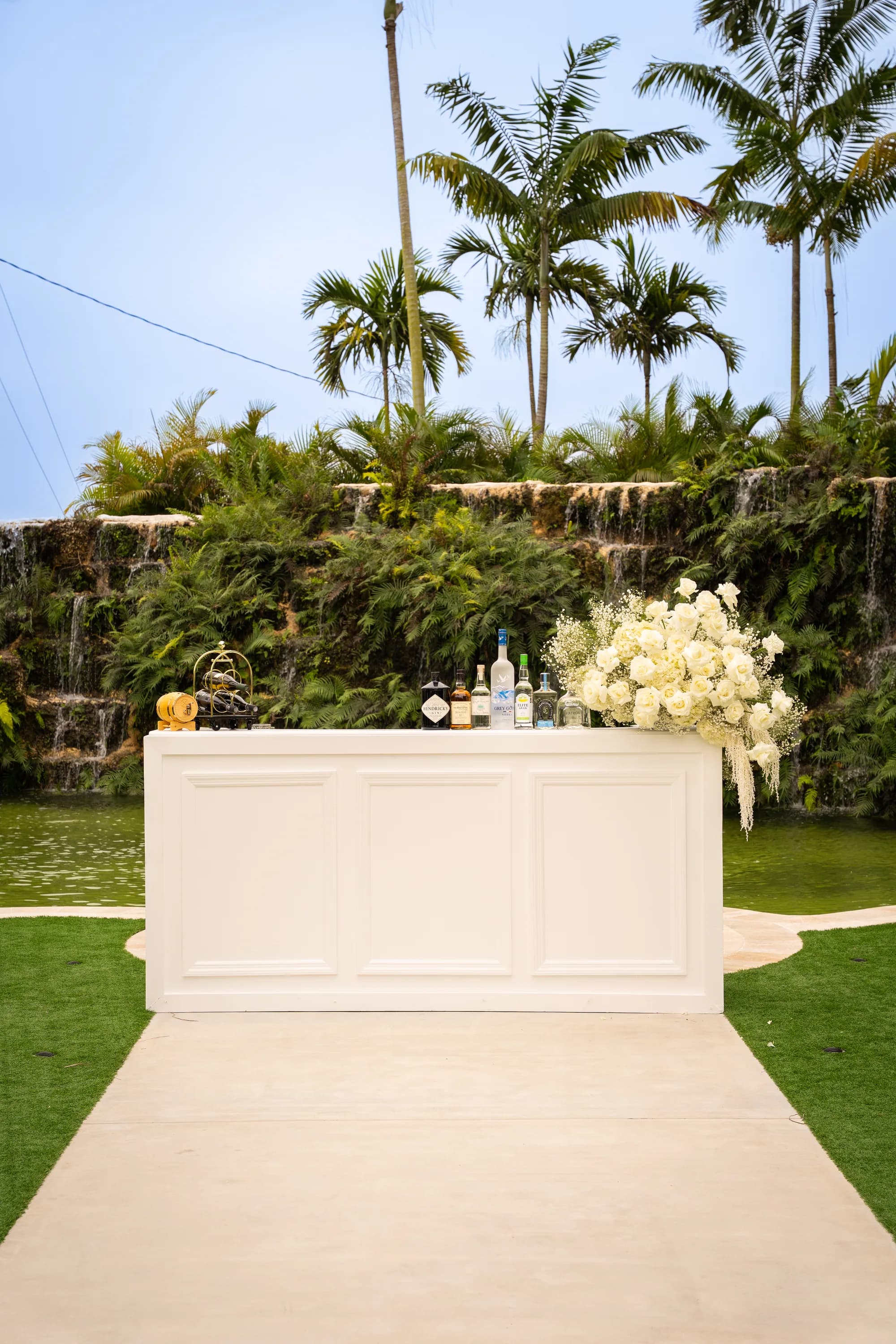 The Salud bar set with spirits and a large white floral arrangement, standing on a walkway in front of a waterfall and lagoon.